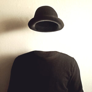 Fotografia che ritrae un maglione nero e un cappello nero che fluttua nell'aria sopra lo stesso, davanti a un muro bianco su cui si proiettano le ombre del maglione e del cappello. Sembra di osservare un uomo invisibile.