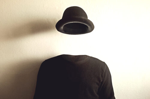 Fotografia che ritrae un maglione nero e un cappello nero che fluttua nell'aria sopra lo stesso, davanti a un muro bianco su cui si proiettano le ombre del maglione e del cappello. Sembra di osservare un uomo invisibile.