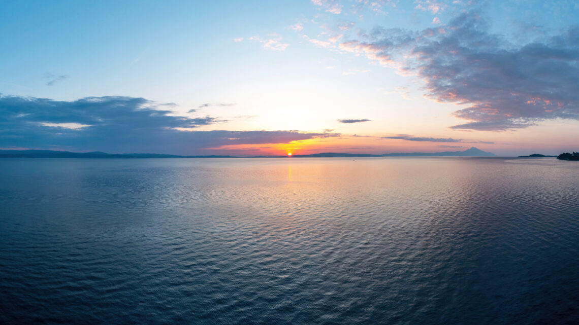 Una fotografia che ritrae un'alba sul mare: il sole, un piccolo disco giallo appena sopra l'orizzonte, spunta da basse montagne in lontananza, in un cielo azzurro chiarissimo intorno al sole, che si fa via via un po' più scuro quanto più ne è distante. Ci sono nuvolette bianche rosa e grigie ai lati dell'inquadratura. Davanti a tutto c'è un mare che riflette i colori del sole e del cielo. Potrebbe essere un arcipelago, o un golfo.