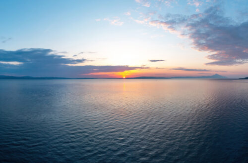 Una fotografia che ritrae un'alba sul mare: il sole, un piccolo disco giallo appena sopra l'orizzonte, spunta da basse montagne in lontananza, in un cielo azzurro chiarissimo intorno al sole, che si fa via via un po' più scuro quanto più ne è distante. Ci sono nuvolette bianche rosa e grigie ai lati dell'inquadratura. Davanti a tutto c'è un mare che riflette i colori del sole e del cielo. Potrebbe essere un arcipelago, o un golfo.