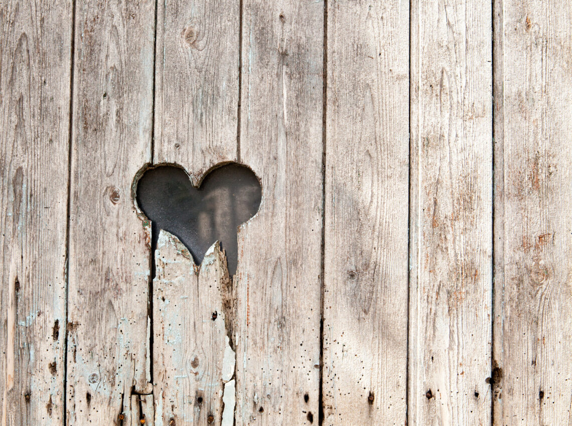 Una foto che ritrae una palizzata di legno consunto con un buco a forma di cuore dai contorni scheggiati a metà altezza, spostato sulla sinistra.