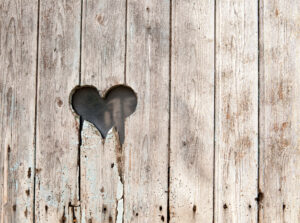Una foto che ritrae una palizzata di legno consunto con un buco a forma di cuore dai contorni scheggiati a metà altezza, spostato sulla sinistra.