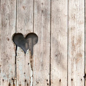 Una foto che ritrae una palizzata di legno consunto con un buco a forma di cuore dai contorni scheggiati a metà altezza, spostato sulla sinistra.
