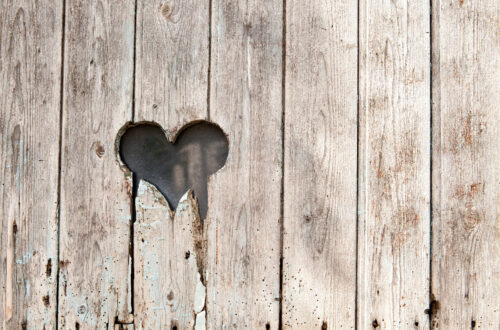 Una foto che ritrae una palizzata di legno consunto con un buco a forma di cuore dai contorni scheggiati a metà altezza, spostato sulla sinistra.