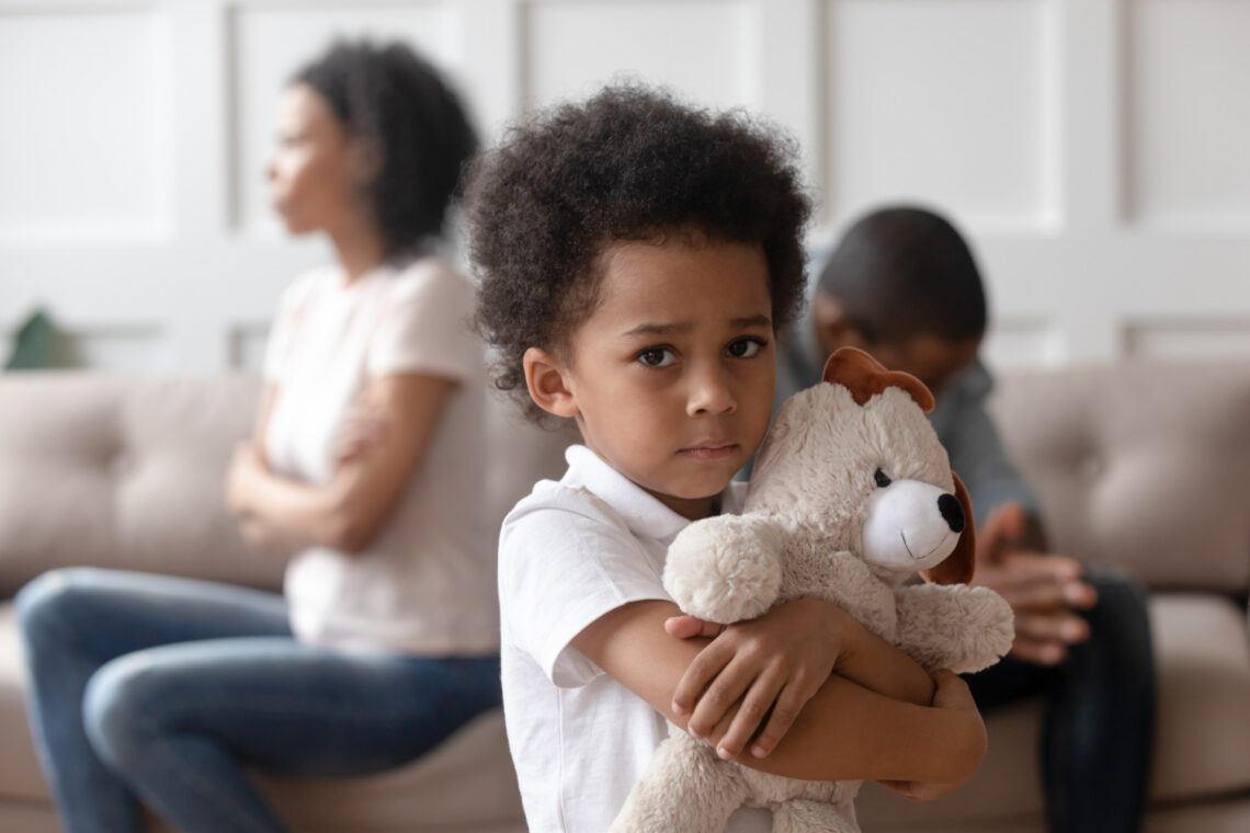 Fotografia che ritrae un bimbo di origini africane che abbraccia con espressione triste il proprio orsetto di peluche bianco mentre sullo sfondo, fuori fuoco, i suoi genitori, seduti su un divano bianco, si danno le spalle, arrabbiati l’uno con l’altra.