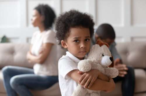 Fotografia che ritrae un bimbo di origini africane che abbraccia con espressione triste il proprio orsetto di peluche bianco mentre sullo sfondo, fuori fuoco, i suoi genitori, seduti su un divano bianco, si danno le spalle, arrabbiati l’uno con l’altra.