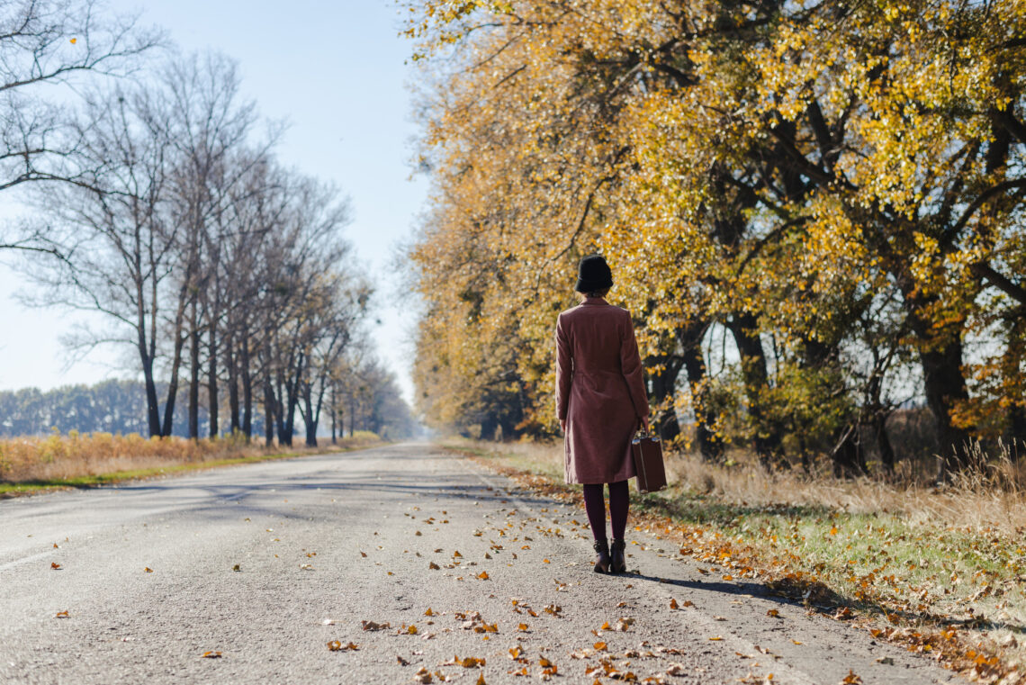 Una foto che ritrae, da dietro, una donna in cammino sul ciglio destro di una strada vuota. La donna indossa un soprabito di velluto, lilla, che le arriva alle ginocchia, e ha un cappellino nero a fungo sulla testa. Sull'asfalto della parte destra della strada ci sono molte foglie gialle e rosse cadute dagli alberi alla destra della strada, ancora frondosi, mentre quelli sulla sinistra sono già spogli e proiettano le loro ombre sull'asfalto. All'orizzonte c'è un cielo azzurro chiaro.