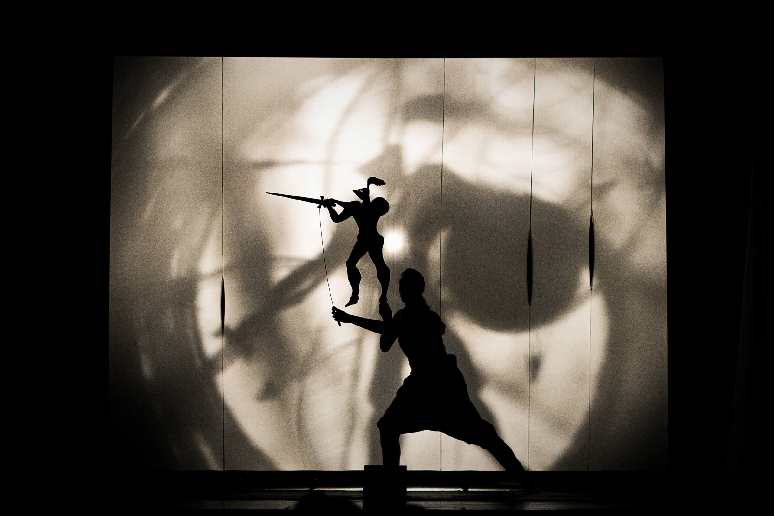 Una foto di scena dallo spettacolo teatrale "Il cavaliere inesistente", basato sull'omonimo romanzo di Italo Calvino. Davanti a un telo retroilluminato, in primo piano c'è la sagoma in ombra di una donna reale che regge una sagoma di cartone raffigurante l'armatura vuota del cavaliere inesistente che brandisce una spada.