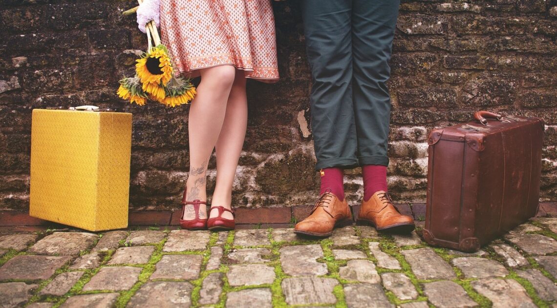 Fotografia che ritrae un muro di mattoni consunti a cui sono appoggiati una donna e un uomo dei quali si vedono solo le gambe e i piedi. Al loro fianco, appoggiate per terra, ci sono due valigie.