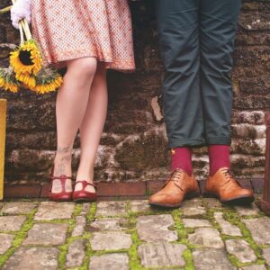 Fotografia che ritrae un muro di mattoni consunti a cui sono appoggiati una donna e un uomo dei quali si vedono solo le gambe e i piedi. Al loro fianco, appoggiate per terra, ci sono due valigie.