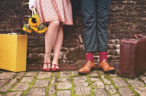 Fotografia che ritrae un muro di mattoni consunti a cui sono appoggiati una donna e un uomo dei quali si vedono solo le gambe e i piedi. Al loro fianco, appoggiate per terra, ci sono due valigie.