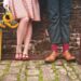 Fotografia che ritrae un muro di mattoni consunti a cui sono appoggiati una donna e un uomo dei quali si vedono solo le gambe e i piedi. Al loro fianco, appoggiate per terra, ci sono due valigie.