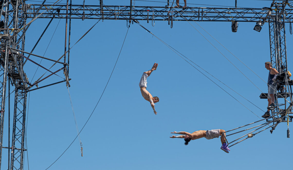 Fotografia che ritrae due trapezisti. Il primo, a sinistra nell'inquadratura, è saltato dal suo trapezio ed è in volo nell'aria con le braccia tese in avanti a cercare la presa del secondo, che a sua volta tende le braccia, appeso a testa in giù con le gambe al suo trapezio. I due sono distanti circa un metro e non si sa se riusciranno a prendersi l'un l'altro.