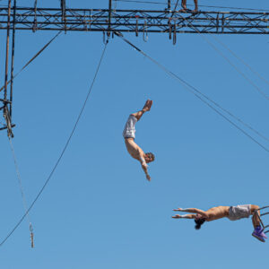 Fotografia che ritrae due trapezisti. Il primo, a sinistra nell'inquadratura, è saltato dal suo trapezio ed è in volo nell'aria con le braccia tese in avanti a cercare la presa del secondo, che a sua volta tende le braccia, appeso a testa in giù con le gambe al suo trapezio. I due sono distanti circa un metro e non si sa se riusciranno a prendersi l'un l'altro.
