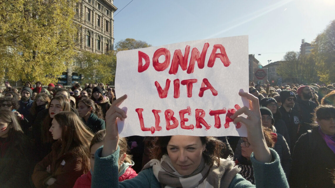 Fotografia di Andrea Delbo per Shutterstock.com che ritrae una piazza, Largo Cairoli a Milano, gremita di persone in manifestazione, per la gran parte donne di tutte le età, ma prevalentemente giovani e adulte, sullo sfondo di alcuni edifici a sinistra, seminascosti da alberi alti e meno alti con foglie di colore verde-giallo, poi un cielo luminoso, dall'azzurro a sinistra al bianco a destra, nei pressi del sole (fuori dall'inquadratura), e altri edifici e piante in fondo a destra, simili a quelli, più vicini, a sinistra. Al centro della folla, in primo piano, c'è una donna adulta, occhi azzurri determinati, volto serio ma con una sfumatura sorridente, capelli lunghi castani tirati indietro, che sostiene sopra la testa, con le braccia in su, un cartello bianco con la scritta “Donna vita libertà” in rosso.