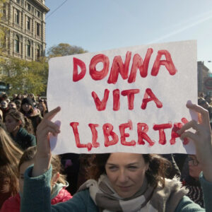 Fotografia di Andrea Delbo per Shutterstock.com che ritrae una piazza, Largo Cairoli a Milano, gremita di persone in manifestazione, per la gran parte donne di tutte le età, ma prevalentemente giovani e adulte, sullo sfondo di alcuni edifici a sinistra, seminascosti da alberi alti e meno alti con foglie di colore verde-giallo, poi un cielo luminoso, dall'azzurro a sinistra al bianco a destra, nei pressi del sole (fuori dall'inquadratura), e altri edifici e piante in fondo a destra, simili a quelli, più vicini, a sinistra. Al centro della folla, in primo piano, c'è una donna adulta, occhi azzurri determinati, volto serio ma con una sfumatura sorridente, capelli lunghi castani tirati indietro, che sostiene sopra la testa, con le braccia in su, un cartello bianco con la scritta “Donna vita libertà” in rosso.