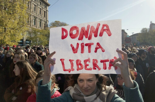 Fotografia di Andrea Delbo per Shutterstock.com che ritrae una piazza, Largo Cairoli a Milano, gremita di persone in manifestazione, per la gran parte donne di tutte le età, ma prevalentemente giovani e adulte, sullo sfondo di alcuni edifici a sinistra, seminascosti da alberi alti e meno alti con foglie di colore verde-giallo, poi un cielo luminoso, dall'azzurro a sinistra al bianco a destra, nei pressi del sole (fuori dall'inquadratura), e altri edifici e piante in fondo a destra, simili a quelli, più vicini, a sinistra. Al centro della folla, in primo piano, c'è una donna adulta, occhi azzurri determinati, volto serio ma con una sfumatura sorridente, capelli lunghi castani tirati indietro, che sostiene sopra la testa, con le braccia in su, un cartello bianco con la scritta “Donna vita libertà” in rosso.