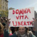 Fotografia di Andrea Delbo per Shutterstock.com che ritrae una piazza, Largo Cairoli a Milano, gremita di persone in manifestazione, per la gran parte donne di tutte le età, ma prevalentemente giovani e adulte, sullo sfondo di alcuni edifici a sinistra, seminascosti da alberi alti e meno alti con foglie di colore verde-giallo, poi un cielo luminoso, dall'azzurro a sinistra al bianco a destra, nei pressi del sole (fuori dall'inquadratura), e altri edifici e piante in fondo a destra, simili a quelli, più vicini, a sinistra. Al centro della folla, in primo piano, c'è una donna adulta, occhi azzurri determinati, volto serio ma con una sfumatura sorridente, capelli lunghi castani tirati indietro, che sostiene sopra la testa, con le braccia in su, un cartello bianco con la scritta “Donna vita libertà” in rosso.
