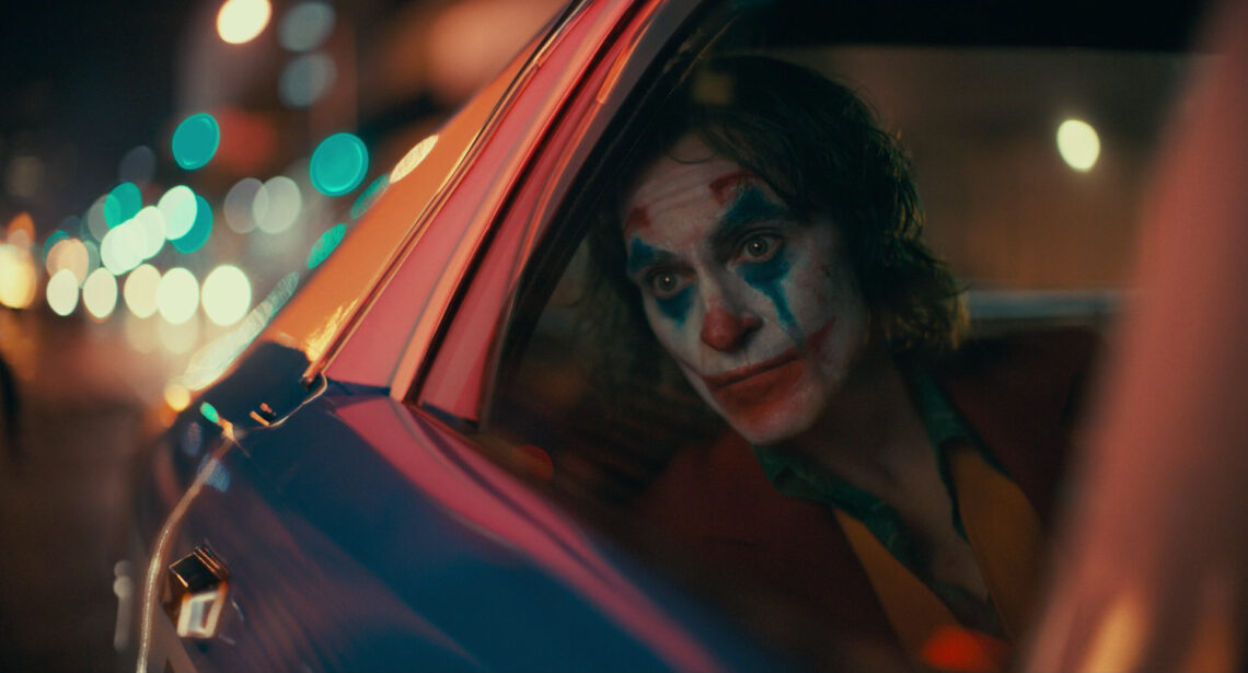 Un fotogramma dal film “Joker” del 2019, di Todd Phillips, con Joaquin Phoenix nella parte del protagonista, Arthur Fleck / Joker, mentre guarda fuori dal finestrino dal sedile posteriore, a destra, di un’auto della polizia, truccato da pagliaccio e con un’aria meditabonda.