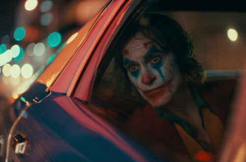 Un fotogramma dal film “Joker” del 2019, di Todd Phillips, con Joaquin Phoenix nella parte del protagonista, Arthur Fleck / Joker, mentre guarda fuori dal finestrino dal sedile posteriore, a destra, di un’auto della polizia, truccato da pagliaccio e con un’aria meditabonda.