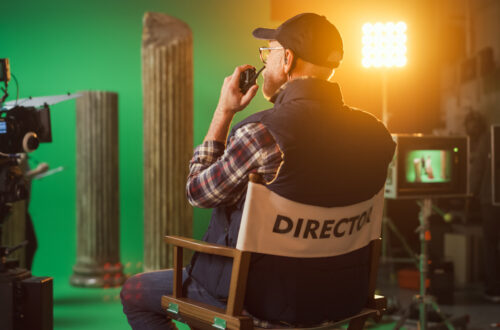 Fotografia che ritrae un set cinematografico con finte colonne greche e uno sfondo verde per il chroma-key. Al centro della scena c’è il regista, di spalle, seduto sulla sua seggiolina, mentre, leggermente voltato verso sinistra, parla con qualcuno attraverso un walkie-talkie che tiene di fianco alla bocca.