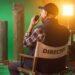 Fotografia che ritrae un set cinematografico con finte colonne greche e uno sfondo verde per il chroma-key. Al centro della scena c’è il regista, di spalle, seduto sulla sua seggiolina, mentre, leggermente voltato verso sinistra, parla con qualcuno attraverso un walkie-talkie che tiene di fianco alla bocca.