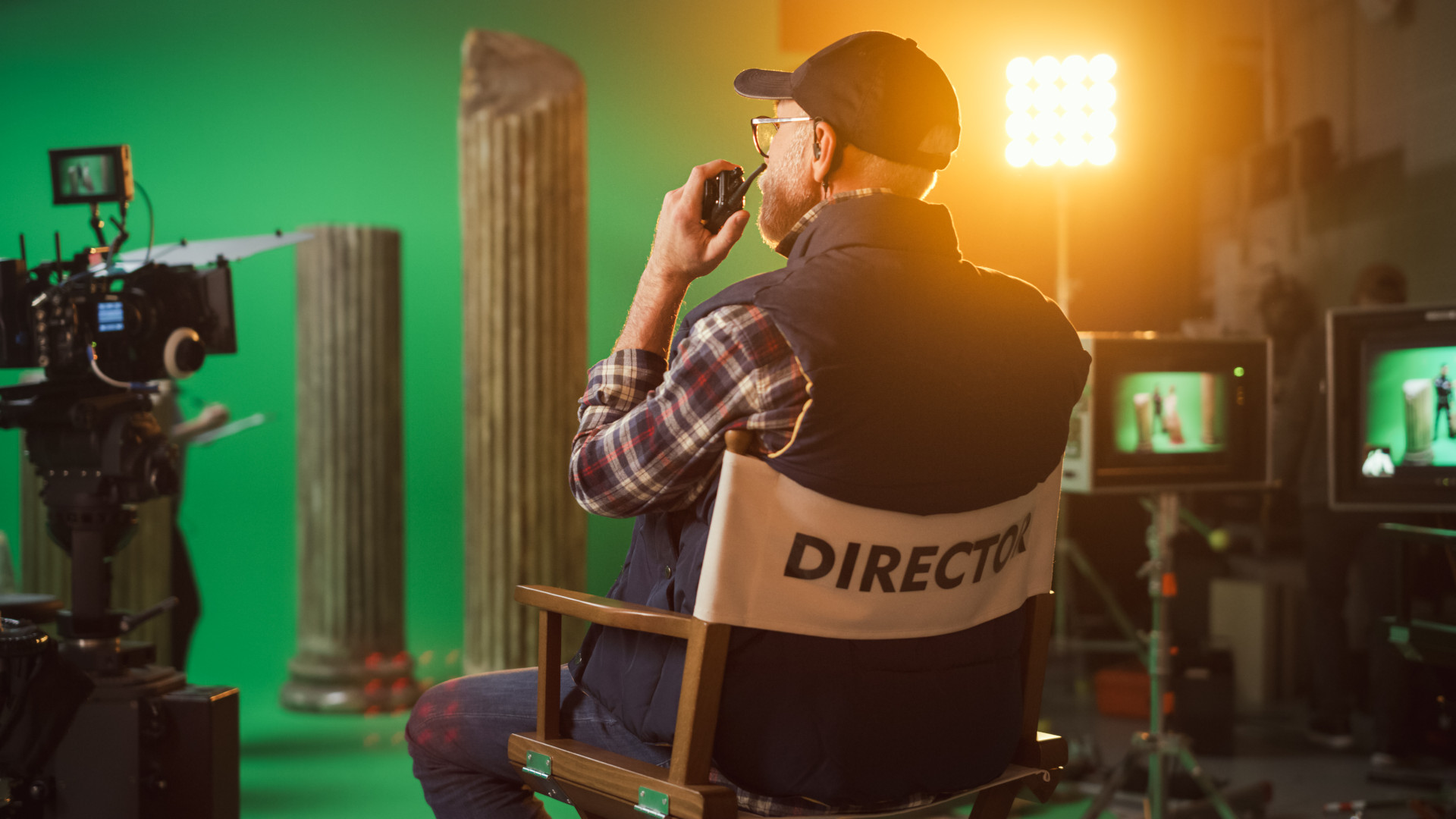 Fotografia che ritrae un set cinematografico con finte colonne greche e uno sfondo verde per il chroma-key. Al centro della scena c’è il regista, di spalle, seduto sulla sua seggiolina, mentre, leggermente voltato verso sinistra, parla con qualcuno attraverso un walkie-talkie che tiene di fianco alla bocca.