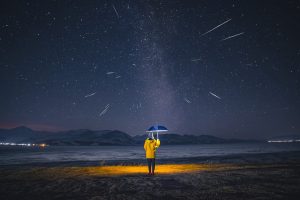 Una foto a colori, in formato 4:3, che ritrae una donna di spalle, in piedi in mezzo a una terra brulla, di notte, che regge un ombrello blu che le copre la testa e che proietta, come una abat jour, una luce calda e circolare su di lei e sul terreno intorno ai suoi piedi. Di fronte alla donna in piedi, all'orizzonte, ci sono delle cime di montagne innevate, e sopra quelle un cielo pieno di stelle dal centro del quale sfrecciano alcune meteore.