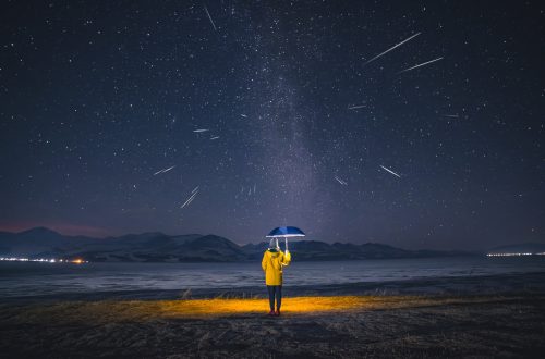 Una foto a colori, in formato 4:3, che ritrae una donna di spalle, in piedi in mezzo a una terra brulla, di notte, che regge un ombrello blu che le copre la testa e che proietta, come una abat jour, una luce calda e circolare su di lei e sul terreno intorno ai suoi piedi. Di fronte alla donna in piedi, all'orizzonte, ci sono delle cime di montagne innevate, e sopra quelle un cielo pieno di stelle dal centro del quale sfrecciano alcune meteore.