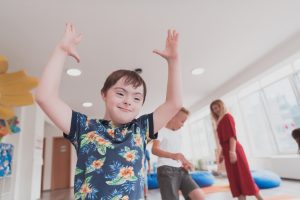 Foto a colori, in formato 16:9, che ritrae un bimbo down sorridente con le braccia alzate in un contesto scolastico