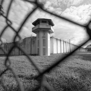 Una foto in bianco e nero che ritrae, attraverso una rete a maglie fuori fuoco, le alte mura perimetrali di cemento bianco di un carcere, sovrastate da filo spinato, con una torre di guardia al centro. Tra la rete e le mura c'è un passaggio ampio di erba. Sullo sfondo, dietro le mura, c'è un cielo nuvoloso.