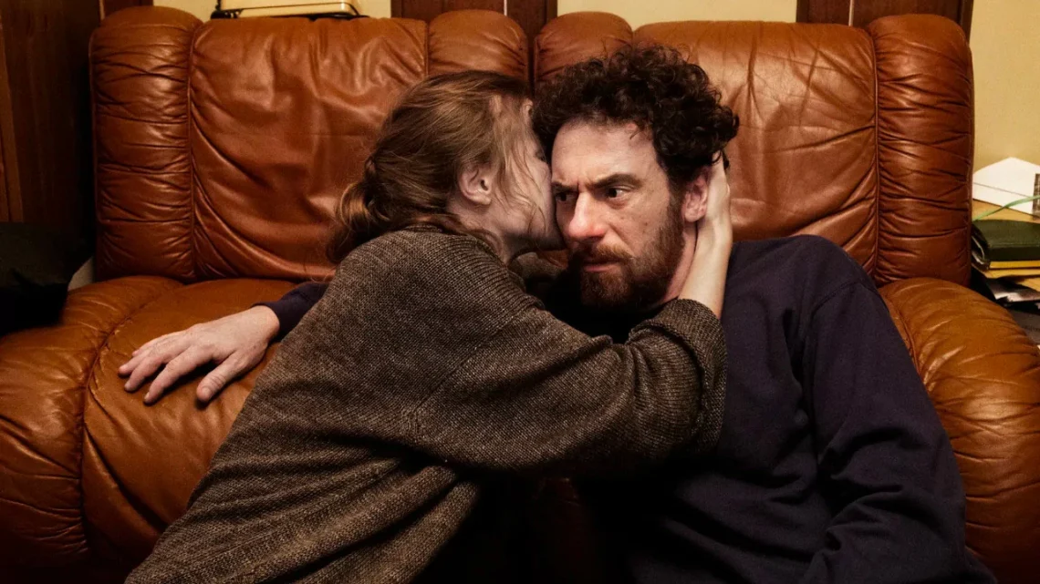 Una scena dal film “Confidenza”, la protagonista sta confidando un suo segreto al protagonista. Sono seduti per terra, ai piedi di un divano, lei ha la mano destra sulla nuca di lui, in un abbraccio che lo tiene vicino, il suo volto non è visibile, mentre è visibile quello di lui, la cui espressione è un po’ intimorita.