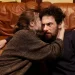 Una scena dal film “Confidenza”, la protagonista sta confidando un suo segreto al protagonista. Sono seduti per terra, ai piedi di un divano, lei ha la mano destra sulla nuca di lui, in un abbraccio che lo tiene vicino, il suo volto non è visibile, mentre è visibile quello di lui, la cui espressione è un po’ intimorita.