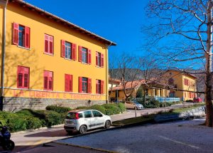 Fotografia a colori, in formato paesaggio, che ritrae la facciata illuminata dalla luce mattutina di un edificio giallo su due piani con tante finestre dagli infissi rossi, alcune chiuse, altre no, e una scritta rossa che recita «La libertà è terapeutica» sulla facciata, sotto la fila di finestre più bassa, quella del primo piano. Davanti all’edificio c’è una strada con alcune automobili posteggiate, e ai suoi fianchi delle siepi e qualche albero. Alla destra dell’edificio ce n’è un altro simile al primo. Sullo sfondo c’è un paesaggio montano sotto un cielo azzurro e terso.