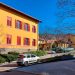 Fotografia a colori, in formato paesaggio, che ritrae la facciata illuminata dalla luce mattutina di un edificio giallo su due piani con tante finestre dagli infissi rossi, alcune chiuse, altre no, e una scritta rossa che recita «La libertà è terapeutica» sulla facciata, sotto la fila di finestre più bassa, quella del primo piano. Davanti all’edificio c’è una strada con alcune automobili posteggiate, e ai suoi fianchi delle siepi e qualche albero. Alla destra dell’edificio ce n’è un altro simile al primo. Sullo sfondo c’è un paesaggio montano sotto un cielo azzurro e terso.
