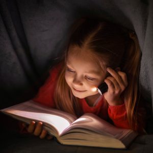 Una fotografia che ritrae una bambina mentre legge un libro alla luce di una piccola torcia elettrica sotto una coperta.