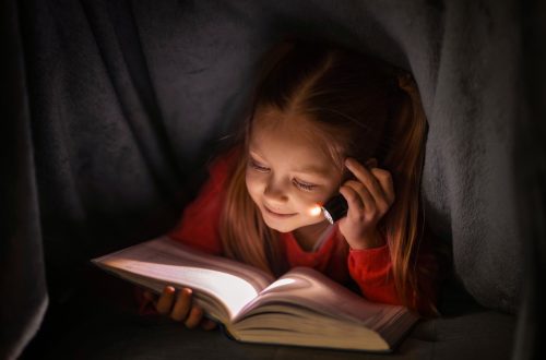 Una fotografia che ritrae una bambina mentre legge un libro alla luce di una piccola torcia elettrica sotto una coperta.