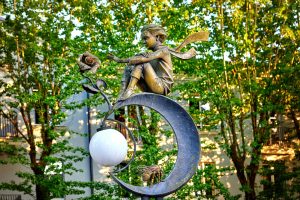 Una foto a colori, in formato orizzontale, che ritrae una scultura in metallo che raffigura il Piccolo Principe, con una lunga sciarpa che sventola dietro la nuca, seduto su una luna con una rosa davanti verso la quale allunga la mano sinistra. La luna è uno spicchio, sulla parte inferiore sorregge inoltre un’ape e tra le due punte dello spicchio una lampada rotonda e bianca. Si trova a Brest, in Bielorussia.