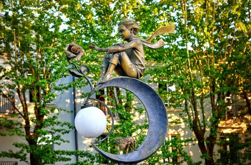 Una foto a colori, in formato orizzontale, che ritrae una scultura in metallo che raffigura il Piccolo Principe, con una lunga sciarpa che sventola dietro la nuca, seduto su una luna con una rosa davanti verso la quale allunga la mano sinistra. La luna è uno spicchio, sulla parte inferiore sorregge inoltre un’ape e tra le due punte dello spicchio una lampada rotonda e bianca. Si trova a Brest, in Bielorussia.