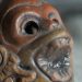 Una foto a colori, in formato panoramico, che ritrae da vicino un fischietto funebre azteco. Sembra essere di terracotta rossa, ha le sembianze di una testa di scimmia con le orecchie decorate da monili di forma prevalentemente circolare, la bocca spalancata in un urlo, il naso piccolo e due grandi occhi vuoti.