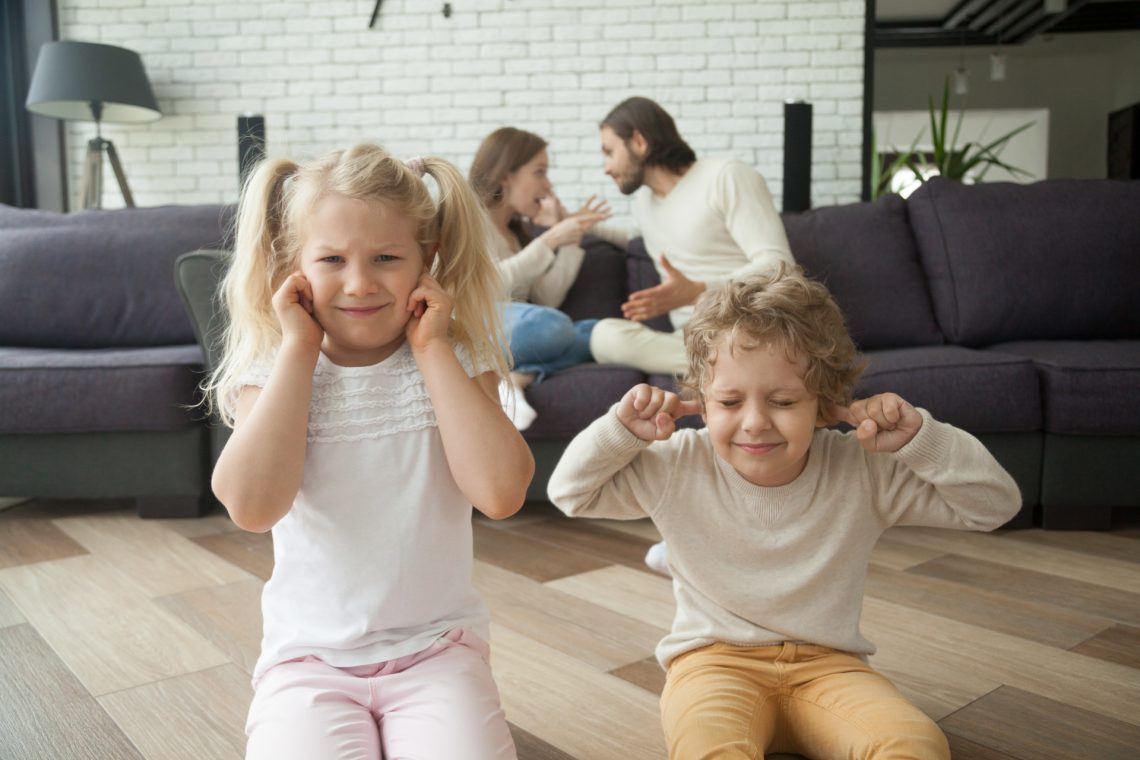 Una fotografia a colori, in formato orizzontale, che ritrae al centro, in primo piano e a fuoco, due bambini, una femmina e un maschio un po' più giovane, seduti in ginocchio sul parquet del pavimento di una bella casa, entrambi con un'espressione infastidita e le dita nelle orecchie per non sentire le parole dell'animata discussione che i genitori, un maschio e una femmina seduti su un divano dietro i bambini, a qualche metro di distanza da loro, un po' fuori fuoco, stanno avendo.