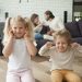 Una fotografia a colori, in formato orizzontale, che ritrae al centro, in primo piano e a fuoco, due bambini, una femmina e un maschio un po' più giovane, seduti in ginocchio sul parquet del pavimento di una bella casa, entrambi con un'espressione infastidita e le dita nelle orecchie per non sentire le parole dell'animata discussione che i genitori, un maschio e una femmina seduti su un divano dietro i bambini, a qualche metro di distanza da loro, un po' fuori fuoco, stanno avendo.