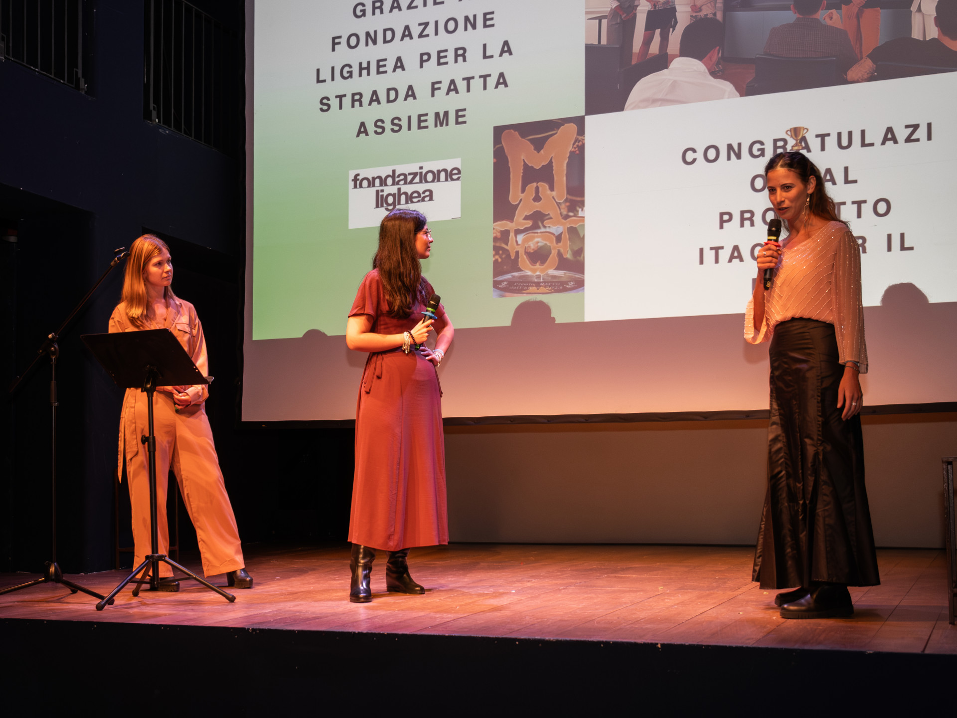 Una foto a colori, in formato orizzontale (4:3), che ritrae un palco sopra il quale ci sono tre giovani donne davanti a uno schermo su cui è proiettata un'immagine da una presentazione. La donna più a destra nell'inquadratura sta parlando in un microfono.
