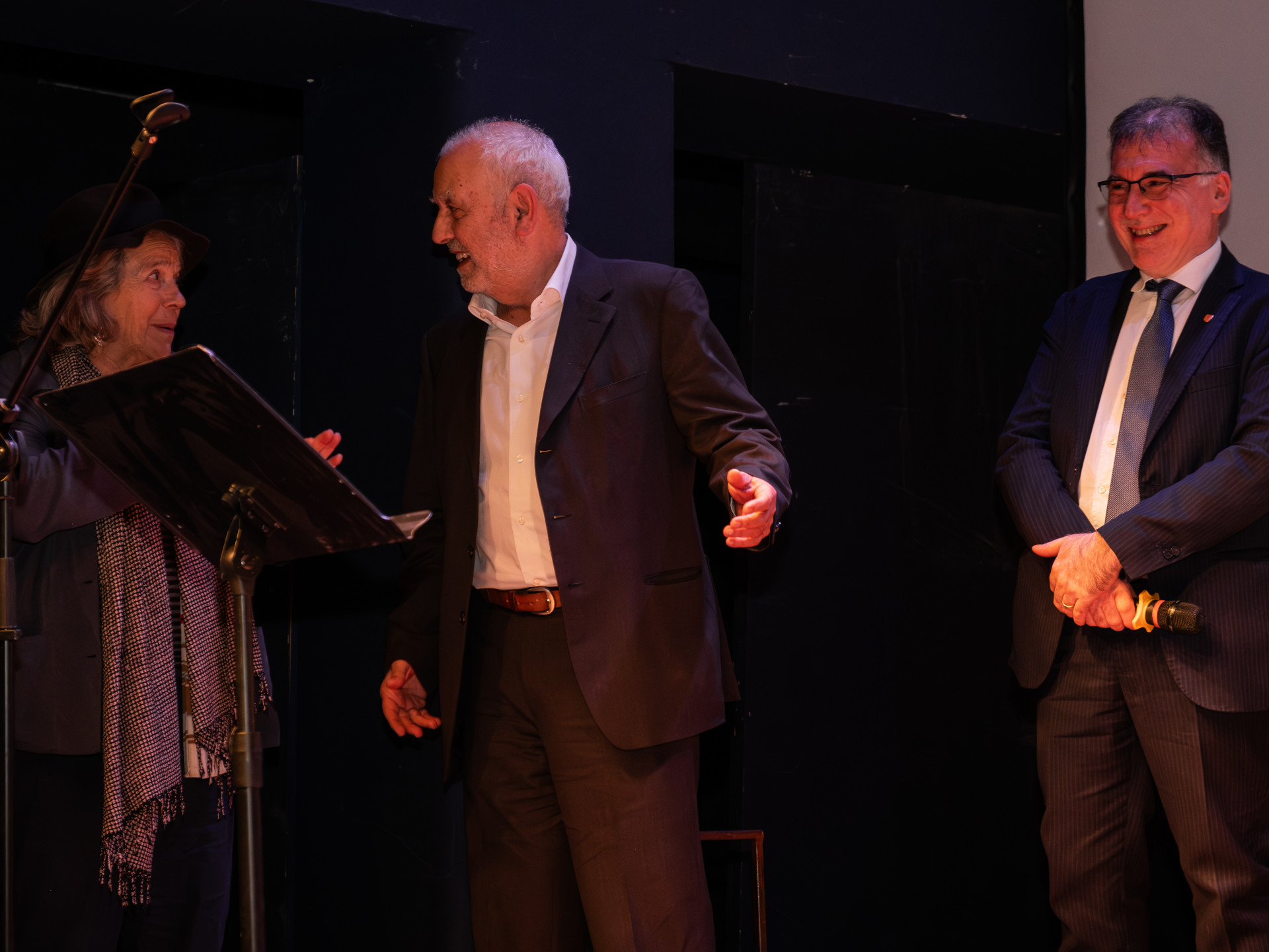 Una foto a colori, in formato orizzontale (4:3), che ritrae un palco sopra il quale ci sono tre persone: una donna a sinistra, di profilo, con la testa rivolta a destra; un uomo al centro, di profilo, con la testa rivolta a sinistra: la donna e l'uomo di stanno guardando e sorridono. A destra c'è un altro uomo, in piedi, con le mani incrociate sulla parte inferiore della pancia; guarda le altre due persone al suo fianco e sorride anch'egli.