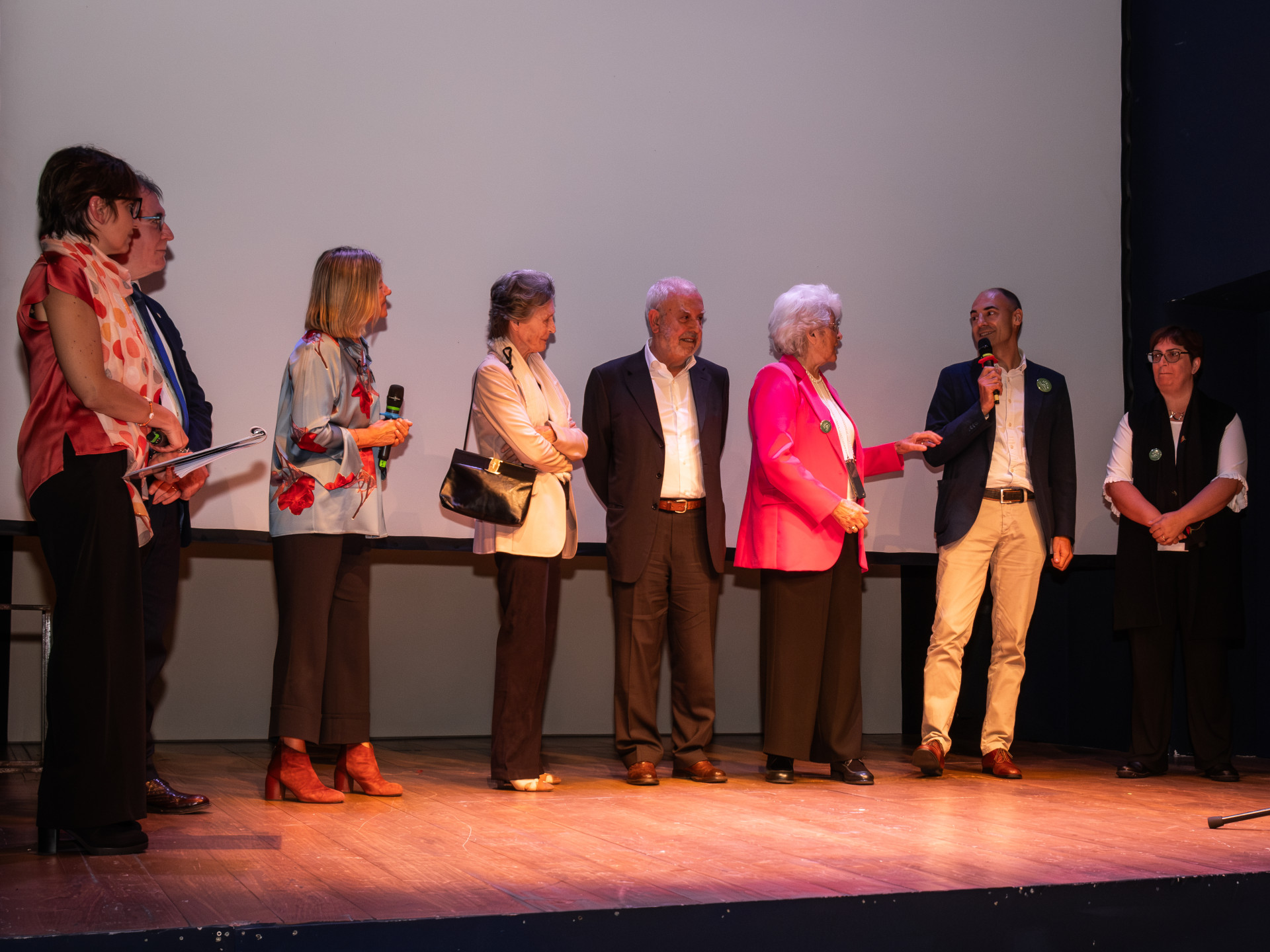 Una foto a colori, in formato orizzontale (4:3), che ritrae un palco sopra il quale, davanti a uno schermo bianco, ci sono otto persone. La sesta, una donna anziana, e l'ottava, un uomo, stanno parlando tra loro, mentre le altre persone li stanno ascoltando, alcune sorridendo.