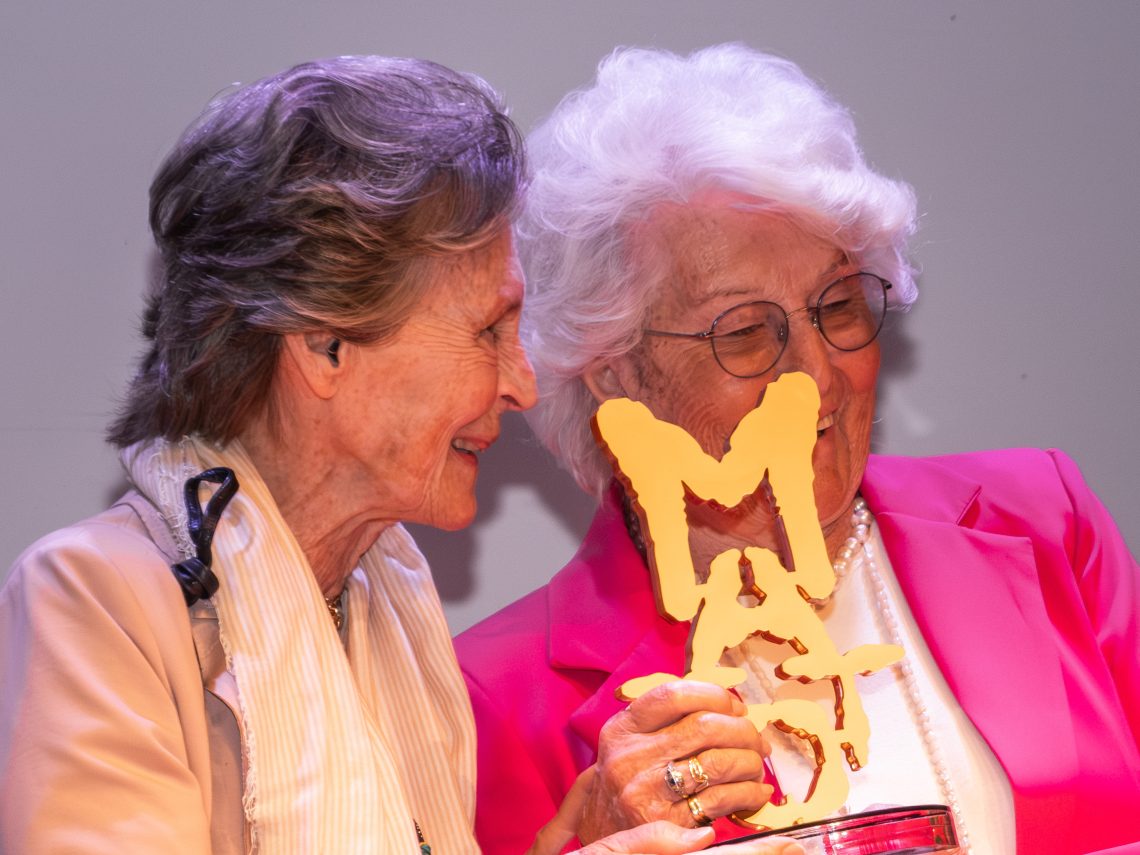 Una foto a colori, in formato orizzontale (4:3), che ritrae due donne anziane, rappresentanti dell’organizzazione “Progetto Itaca”, mentre ritirano il premio “Matto dell’anno” 2024, una piccola scultura che raffigura la scritta “Matto” in caratteri irregolari e in verticale. Le due donne sono in piedi e sorridono.