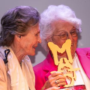 Una foto a colori, in formato orizzontale (4:3), che ritrae due donne anziane, rappresentanti dell’organizzazione “Progetto Itaca”, mentre ritirano il premio “Matto dell’anno” 2024, una piccola scultura che raffigura la scritta “Matto” in caratteri irregolari e in verticale. Le due donne sono in piedi e sorridono.