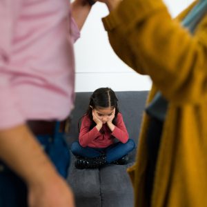 Una foto a colori, in formato orizzontale, che ritrae sul lato sinistro e destro, in primo piano e fuori fuoco, di profilo, un uomo e una donna che si fronteggiano animatamente, dei quali si vedono solo i busti. Nello spazio tra i due busti, più lontana, c'è una bimba seduta a gambe incrociate su un divano grigio, che si tiene il viso tra le mani con la testa un po' reclinata in avanti e un'espressione triste.
