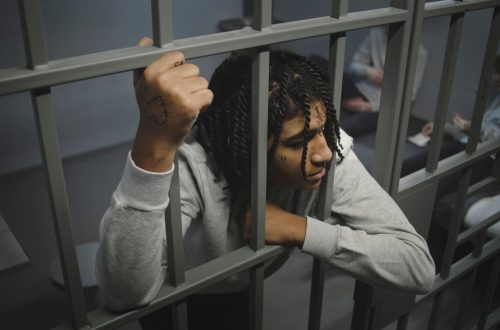 Una foto che ritrae un ragazzo africano, con treccine in testa e qualche tatuaggio sul viso e sul dorso della mano destra, che guarda con espressione molto seria e assorta fuori dalla cella in cui è rinchiuso, affacciato alle sbarre del suo cancello, mentre alcuni compagni di cella, sullo sfondo, giocano a carte seduti sopra un letto.