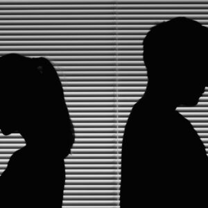 Una foto in bianco e nero che ritrae le silhouette nere di una donna e un uomo, entrambi di profilo, che si danno le spalle davanti a una veneziana chiusa da cui entra poca luce.